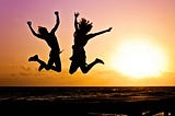 Two people jumping into the air in front of the sunset and the ocean