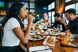 People eating and smiling, seated at a table with many different plates of food and drinks.