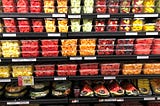 Supermarket shelves filled with overpriced plastic containers of pre-cut watermelon, pineapple, melon, and assorted fruit.
