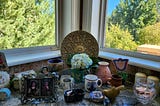 Knick knacks including an owl mug with a hydrangea blossom in front of two windows on a kitchen ledge.