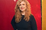 Photo: Mindy Tucker. Woman with long red hair, wearing a navy sweater and navy pants. The early-autumn sun shines through trees visible above the red and gold backdrop.