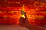 Against a backgroud red with yellow hearts, a butterfly perches on a person’s thumb.