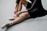 A ballerina dressed in a black leotard and skirt sitting on the floor. She is tying up one her pink ballet pointe shoes.