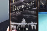 Uma mão segura um exemplar do livro O Demônio na Cidade Branca, de Erik Larson; em segundo plano, há uma casa e uma piscina.