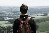 Male with short dark hair facing a countryside in front of him. His back his turned and he he wearing a brown backpack.