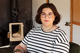 A lady with short dark brown hair wearing glasses and a striped T shirt sits in front of a log-burning stove
