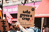 Person holding sign that says: Who gives a crap. Then side comment says “We do.”