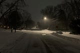 An intersection plowed, but still covered in snow. A single street light shines down on the intersection as night falls.