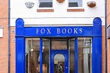 Photograph of the front of the Fox Books bookshop, shows a blue front, with door and windows either side. Red and orange bricks on either side, then a grey roof above. Pavement in front, with a wooden foldable sign