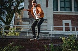 A young man wearing an orange hoodie and toque jumps off a brick wall in a parkour move.