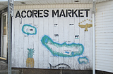A map of several islands of the Azores painted on the side of a small building. Decorating the map are the golden goshawk and stars of the Azorean flag, a whale, a pineapple and some cows.