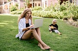 A mother working on a laptop, sitting on grass with her toddler, surrounded with lush plants and trees that cast ample amount of shadow.