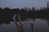 A girl by the lake with a reflection of trees and the city with her hands and feet up in the air.