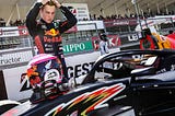 Liam Lawson is running his hands through his hair while contemplating his race helmet, which is resting on the nose of his race car. He’s wearing Red Bull team kit.