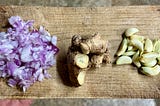 On a wooden tray, chopped onions, ginger, and peeled garlic are placed in individual groups.