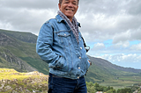 My father wearing a denim jacket and a scarf with Welsh mountains and a cloudy sky as the backdrop.