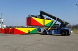A crane lifts a container painted in red, yellow, and green, next to four identical containers.