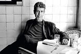 A black & white photo of Anthony Bourdain sitting at a restaurant table.
