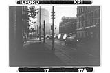 A 1980s urban street, gritty in black and white, cars sit at the light, the sun silhouettes the buildings and passers by