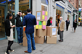 Two researchers standing behind a stack of cardboard boxed with posters reading ‘help us design a better grafton quarter’ in Dublin. Each researcher is talking to a member of the public.