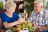 Older woman and man smile while eating a salad.