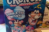 a box of “Captain Crunch Cotton Candy Crunch cereal”, with a bowl of the cereal in front of it. Both are blue and pink.