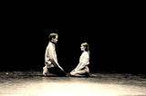 Two actors kneel facing each other on an empty, minimalist stage. They appear calm, focused and open to each other, ready to listen thoughtfully.