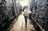 A scene from the movie Labyrinth: A young, white woman in a white poet’s blouse, vest, and jeans runs along a desolate labyrinth hallway with broken bricks and dead vines on an overcast day.