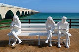 Three white figurines sit on a white bench in front of the ocean, looking at their phones. Two of them sit together on one side of the bench, while the other sits apart from them on the other side.