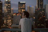 Man in white shirt overlooks city buildings and lights poignantly.