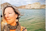 An older woman stares into the distance from a sailboat at anchor, wind-whipped waves and rocky cliffs in the distance.