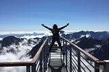 A woman triumphantly stands on a railing viewing the vast cloudy mountains in front of her.