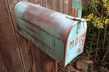 Photo of an old, worn mailbox, green patina or paint, with its flag up, attached to a wooden fence. The word MAIL is on the front flap.