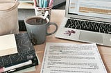 a partially visible laptop kept on a table alongside papers, notebooks, a cup of pens, and a cold espresso.