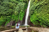 man wearing backpack sitting in front of waterfall surrounded by green vegetation