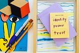 Tableau of a colorful magazine cover featuring an illustration of a hand making a peace sign, an eye, and a heart. A pencil, Sharpie, and paint brush lay on top of the magazine. A post-it with a hand-written list “ identity, power, trust” rest atop a framed piece of colorful line art.