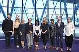 9 people stand on a purple carpet in front of a big window looking out onto the river Thames.