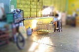 A rattan baby crib sits in a well-lit corner of an open market in Cebu City, Philippines. Sunlight casts a warm glow on the crib, highlighting its simplicity. Nearby, a person sits on a chair, partially visible, while the surrounding area reflects the bustling yet rustic ambiance of the market with scattered items and vibrant colors.