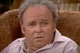 Sitting in his worn living room wing back chair, Carroll O’Connor as Archie Bunker gives one of his signature stares
