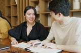 A man and woman chatting over their notebooks at a table in what appears to be a public space, possibly a library