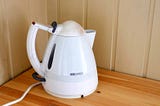 An old, white plastic electric water kettle on a wooden bench