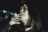 With a black background, a young woman takes up most of the screen. She is holding her lips as she seems to be singing to a microphone that is at the right corner. She has a concerned look but it is unknown what she is looking at.