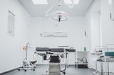 A photo of a surgical room. A dark bed is in the middle, on a hydraulic lift. A large lamp is positioned over the bed. A variety of tables and carts with supplies sit around the bed. The walls are white and the floor is gray.