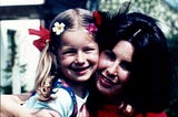 Mother hugging her young daughter who has red bows and flowers in her hair