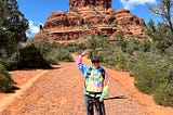 CC in a rainbow hoodie standing in front of Bell Rock red rock vortex in Sedona with a strong arm up