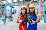 Two young women standing back to back, wearing hard hats and standing in an engineering facility.