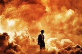 A small silhouette of Cillian Murphy as Dr. Oppenheimer, wearing his suit and hat, against the backdrop of a larger atomic explosion.