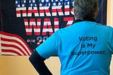 My colleague Lois facing a flag that says “Save Democracy Harris Walz” wearing a blue t-shirt that says “Voting is My Superpower.”