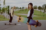 woman with shoulder-length hair holding a rainbow skateboard. another woman skates near her.