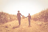 A mother and daughter walk hand in hand along a sunlit path bordered by tall grass, bathed in warm golden light that feels like early autumn.